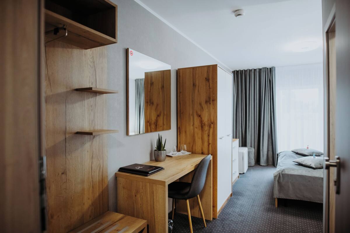 A hotel room with a wooden desk, a chair, a bed with a grey duvet, a small potted plant, and a mirror on the wall.