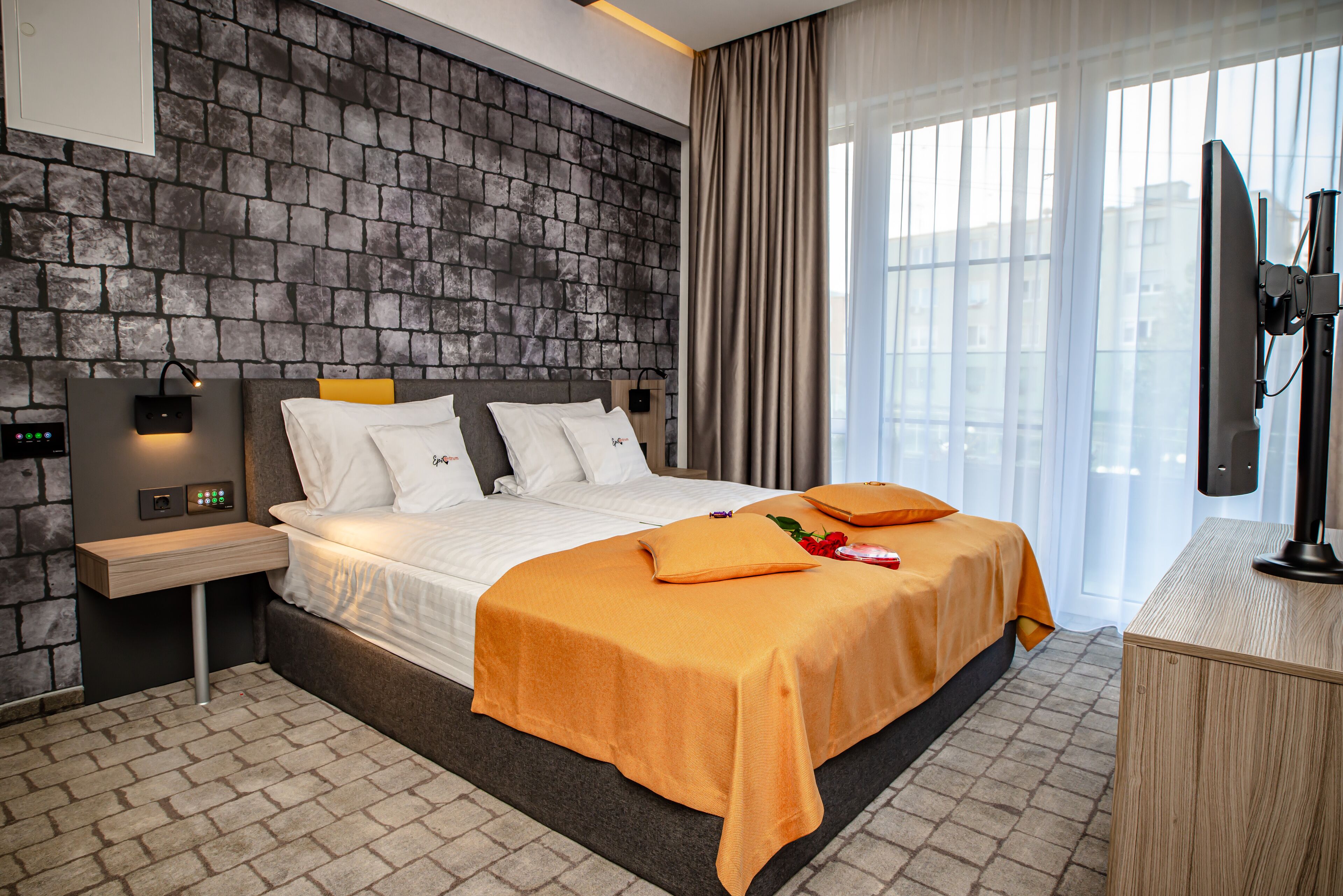 A hotel room with a stone-patterned headboard, a bed with white bedding and orange accents, a bedside table with a lamp, and a television on a wooden stand.