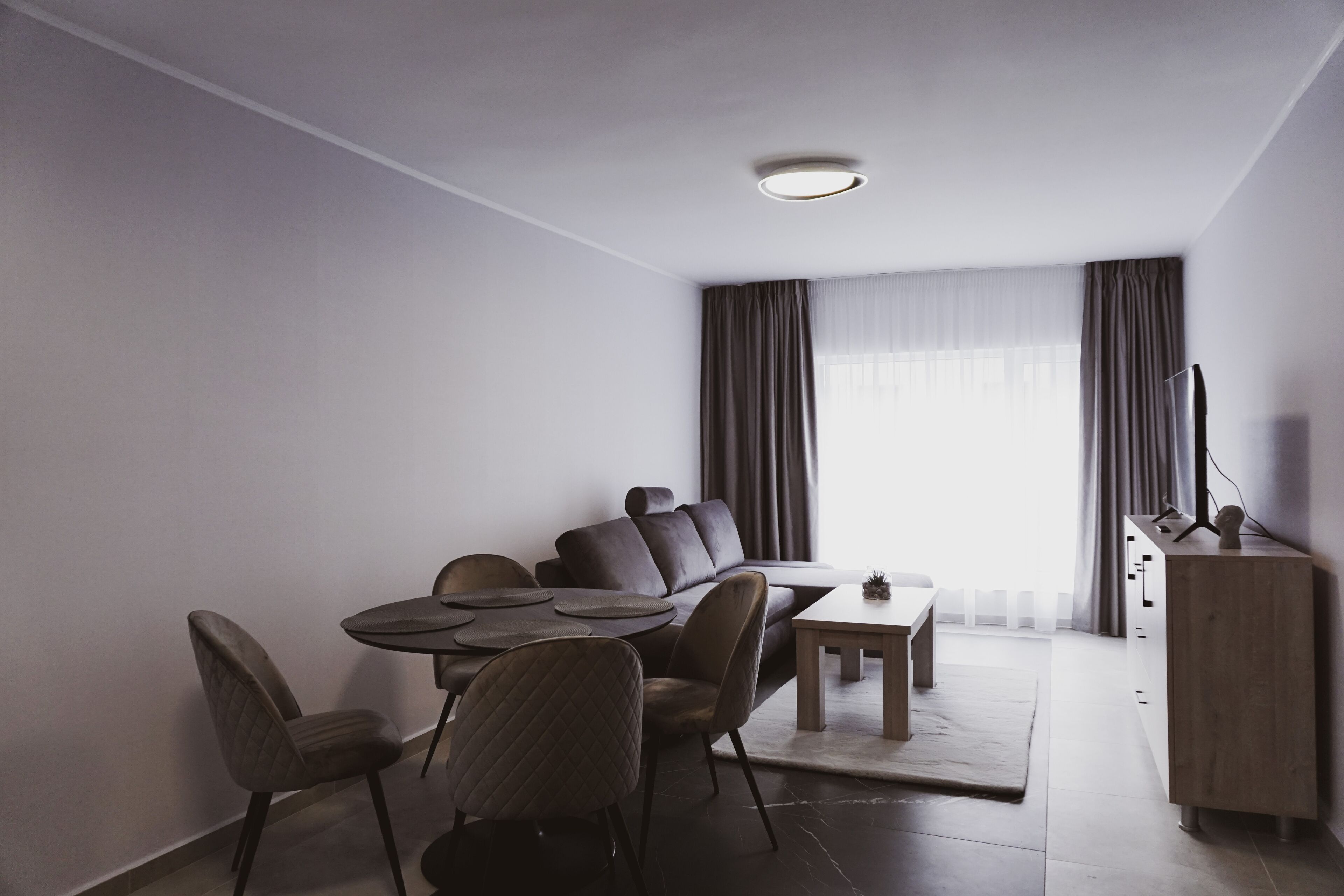 A modern hotel room with a dining area, a sofa, a TV, and a cabinet.