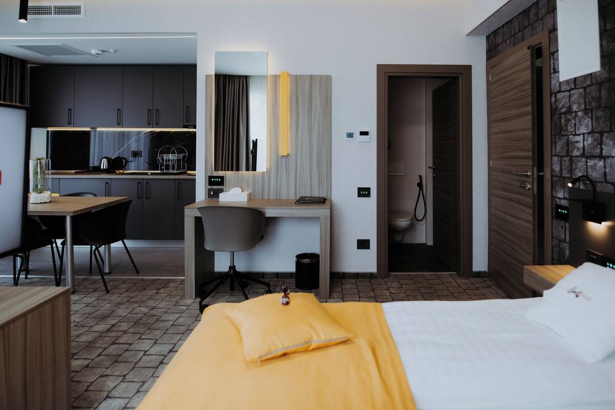 A hotel room with a bed, a desk, a chair, a small table, and a kitchenette in the background.