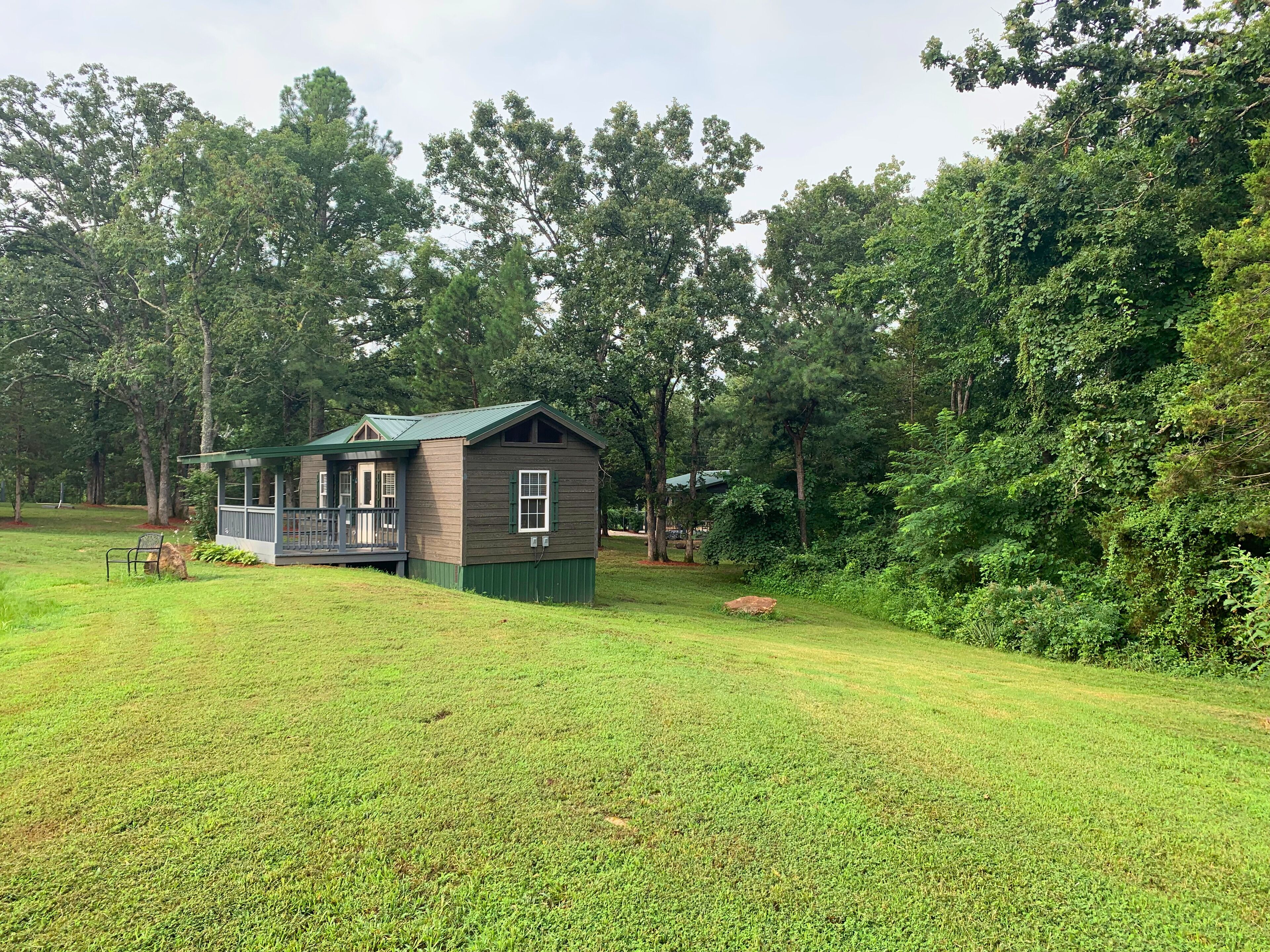 A small, single-story cabin with a porch, surrounded by a grassy field and dense trees.