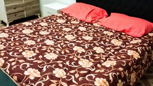 Ein Bett mit floralem Überwurf und zwei roten Kissen, ein Holzschrank und ein weißer Nachttisch.