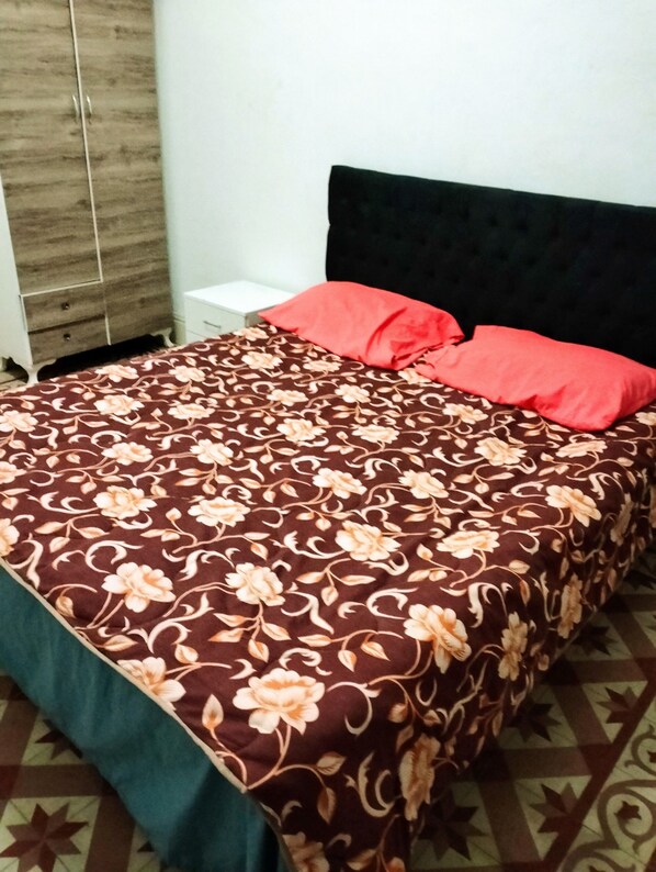 A bed with a floral bedspread and two red pillows, a wooden wardrobe, and a white bedside table.