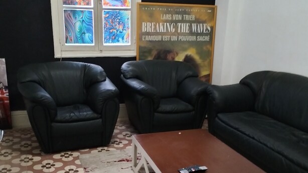 Ein Zimmer mit zwei schwarzen Ledersesseln, einem braunen Couchtisch und einer schwarzen Couch. Auf dem Boden liegt ein Teppich mit Muster und an der Wand hängt ein Poster.