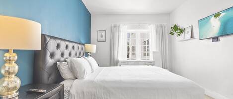 A bedroom with a large bed, a nightstand with a lamp, a TV on the wall, and a window with curtains.