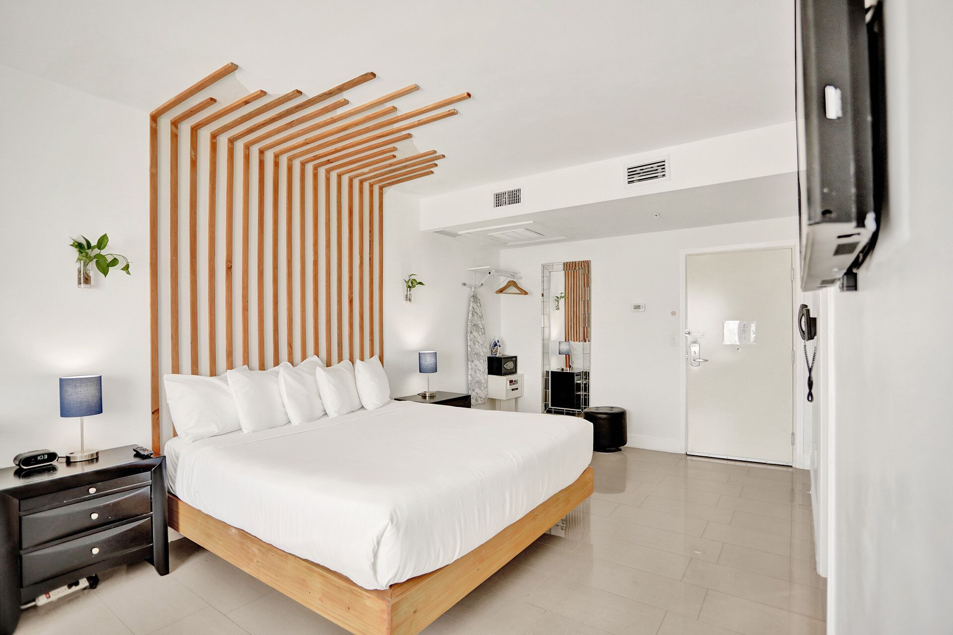 Quarto de hotel moderno com estrutura de cama em madeira, mesa de cabeceira com luminária, uma pequena planta e um espelho.