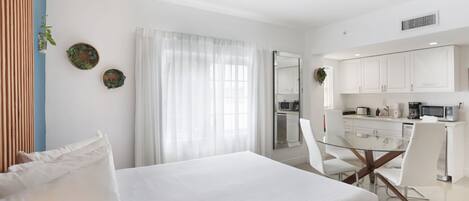 Une chambre d’hôtel moderne avec un grand lit, une table à manger en verre, des murs blancs et un coin cuisine.