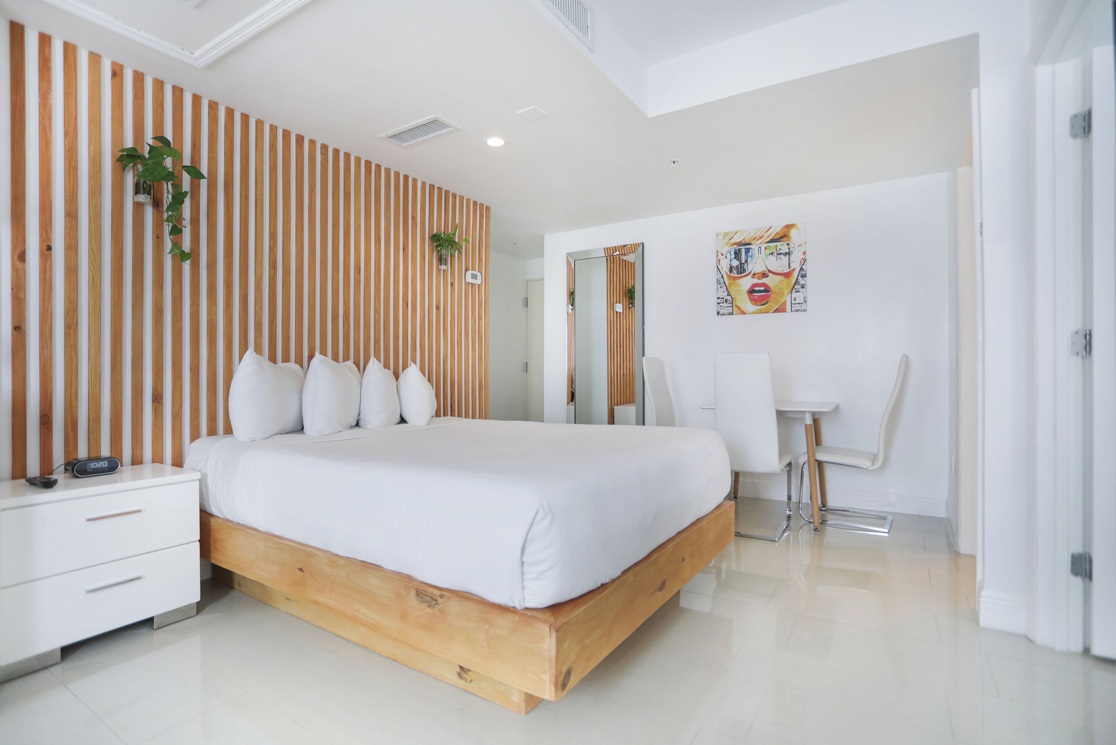 A modern bedroom with a wooden bed frame, white bedding, a bedside table, a chair, and a wall-mounted mirror.