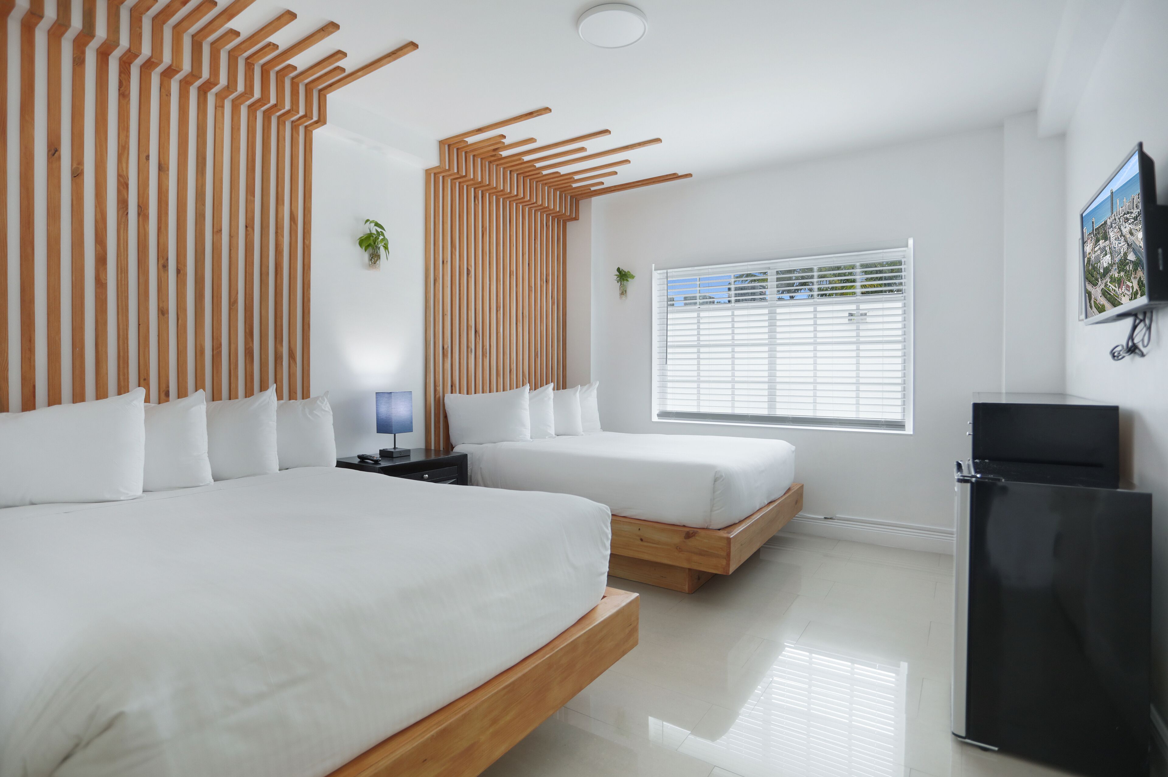 A modern hotel room with two beds, a black refrigerator, a window with blinds, and a mounted TV.