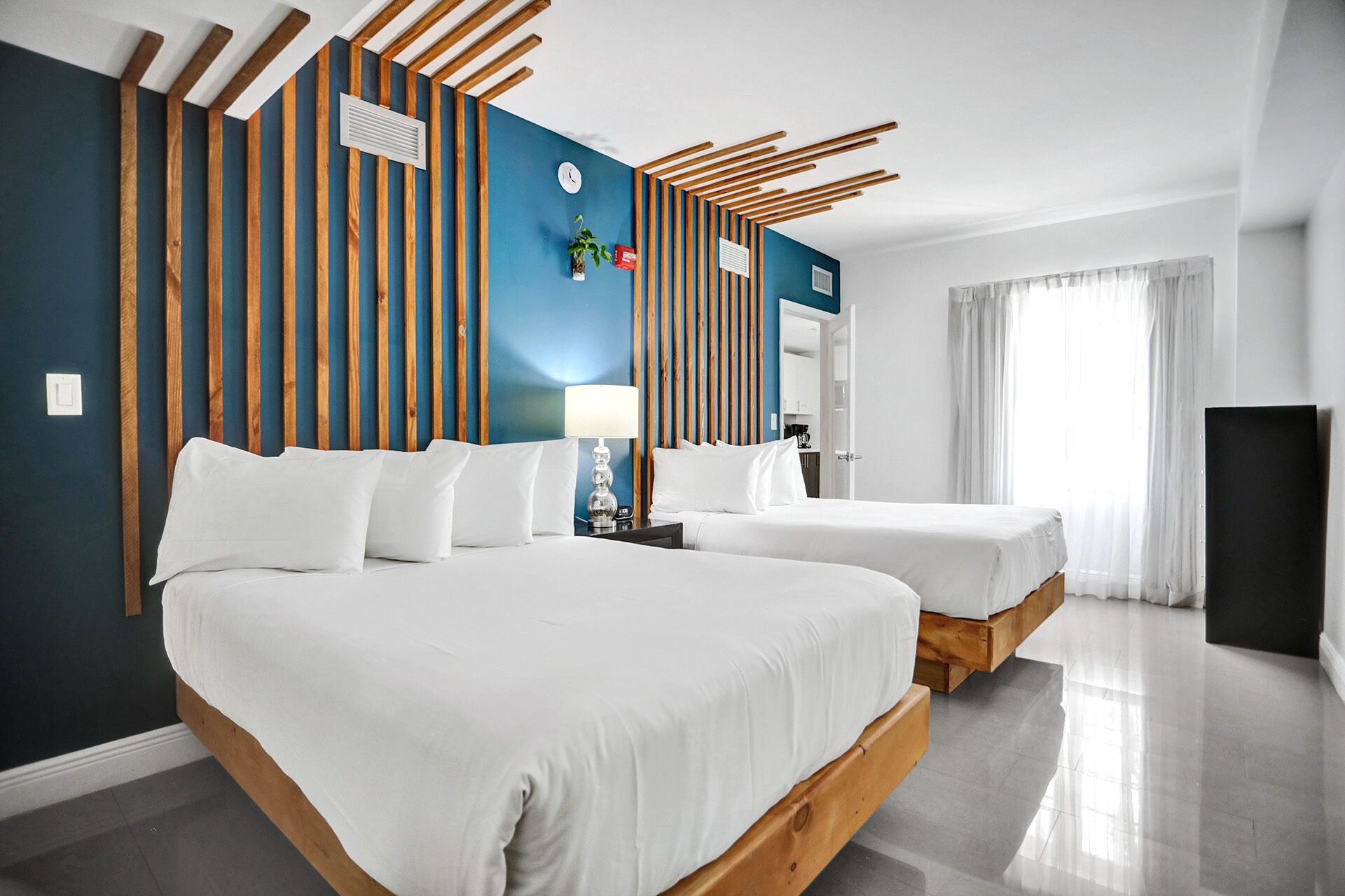 A hotel room with two beds, a wooden headboard, a blue accent wall, a clock, and a window with curtains.