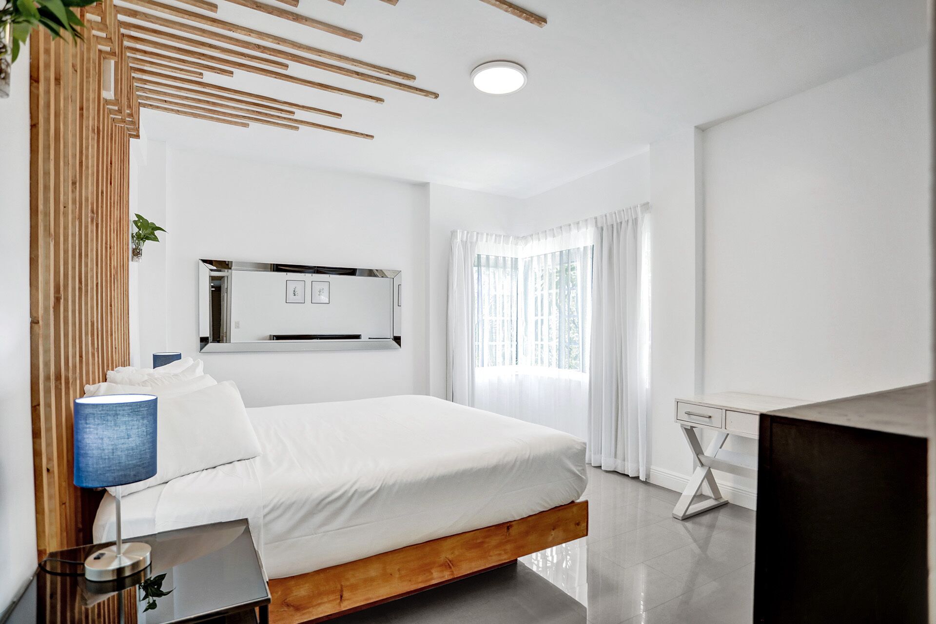 Une chambre moderne avec un cadre de lit en bois, une table de chevet avec une lampe, un grand miroir et une fenêtre avec des rideaux.