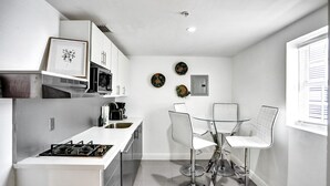 Een kleine keuken met een gasfornuis, magnetron en witte kastjes. Er is een eethoek met een glazen tafel en witte stoelen.