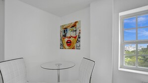 A room with a round glass table and two white chairs with black frames. There is a large painting on the wall and a window with a city view.