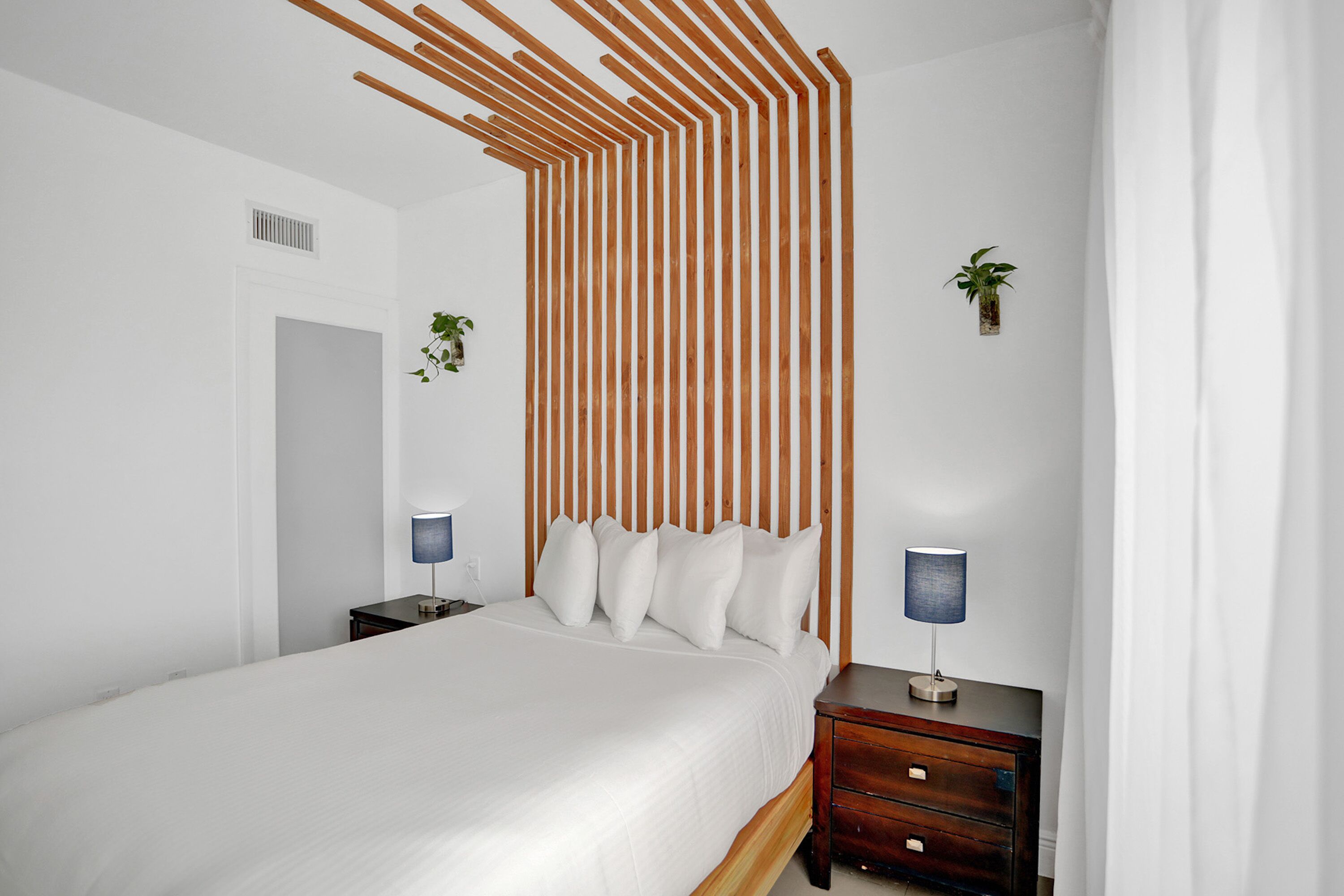 Ein modernes Schlafzimmer mit einem Holzschrankgitter als Kopfteil, einem Bett mit weißen Leinen, zwei Nachttischen mit Lampen und einem kleinen Topf mit Pflanze an der Wand.