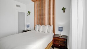 A modern bedroom with a wooden slatted headboard, a bed with white linens, two bedside tables with lamps, and a small potted plant on the wall.