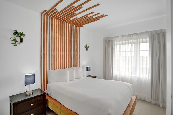 Un dormitorio con una cama, dos mesitas de noche, una mesita auxiliar, una planta en la pared y una ventana con cortinas traslúcidas.