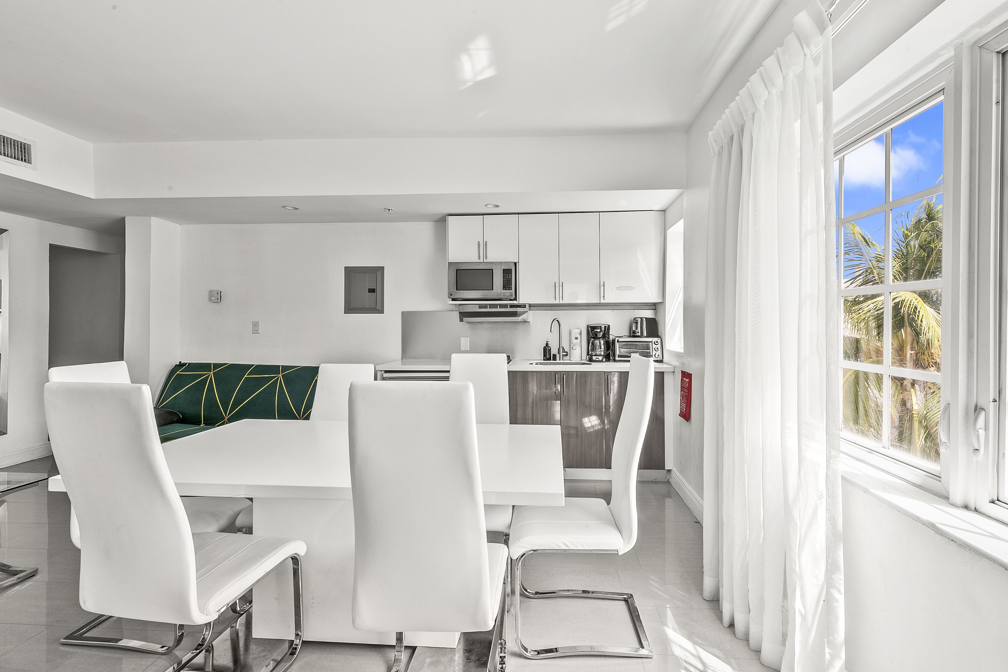 A modern kitchen with white cabinets, a microwave, and a coffee maker. There is a dining area with white chairs and a table.