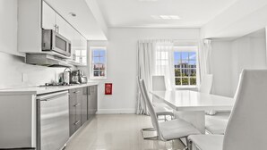 A modern kitchen with stainless steel appliances, a dining area with white chairs, and large windows with curtains.