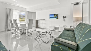 A modern living room with a glass coffee table, a sofa with geometric patterns, a dining area with a white table and chairs, and a large window with a view of a beach.
