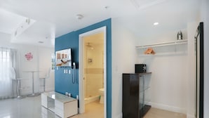 Ein modernes Hotelzimmer mit blauer Akzentwand, einer Glasdusche, einer weißen Toilette, einem schwarzen Kühlschrank und einem kleinen Tisch mit einem Stuhl.