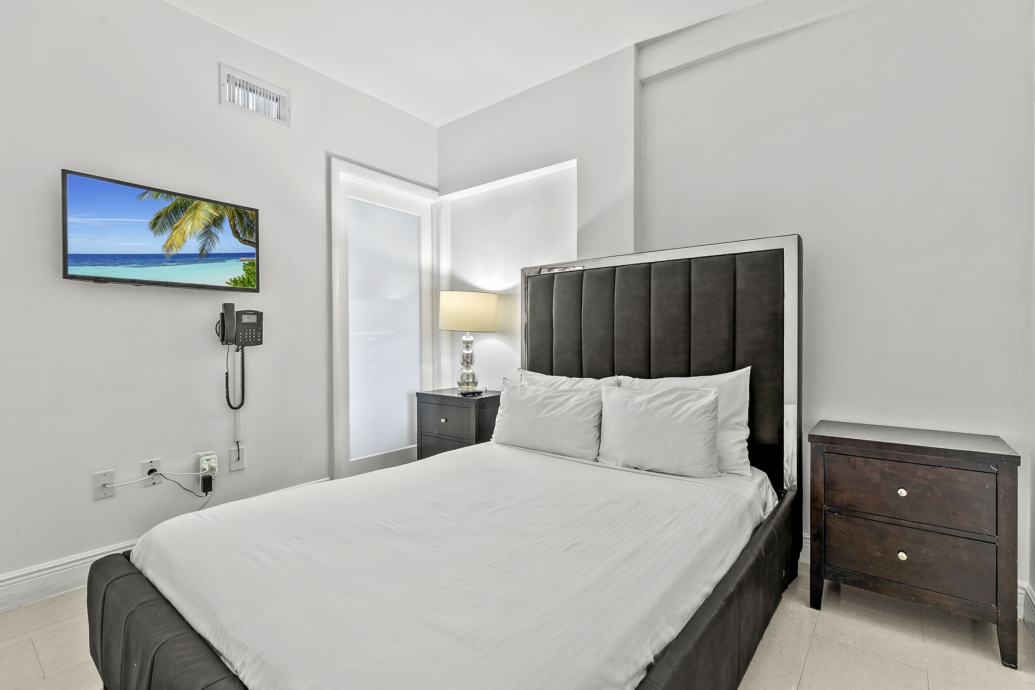 A neatly arranged bedroom with a bed, bedside tables, a nightstand, a lamp, a phone, and a TV mounted on the wall.