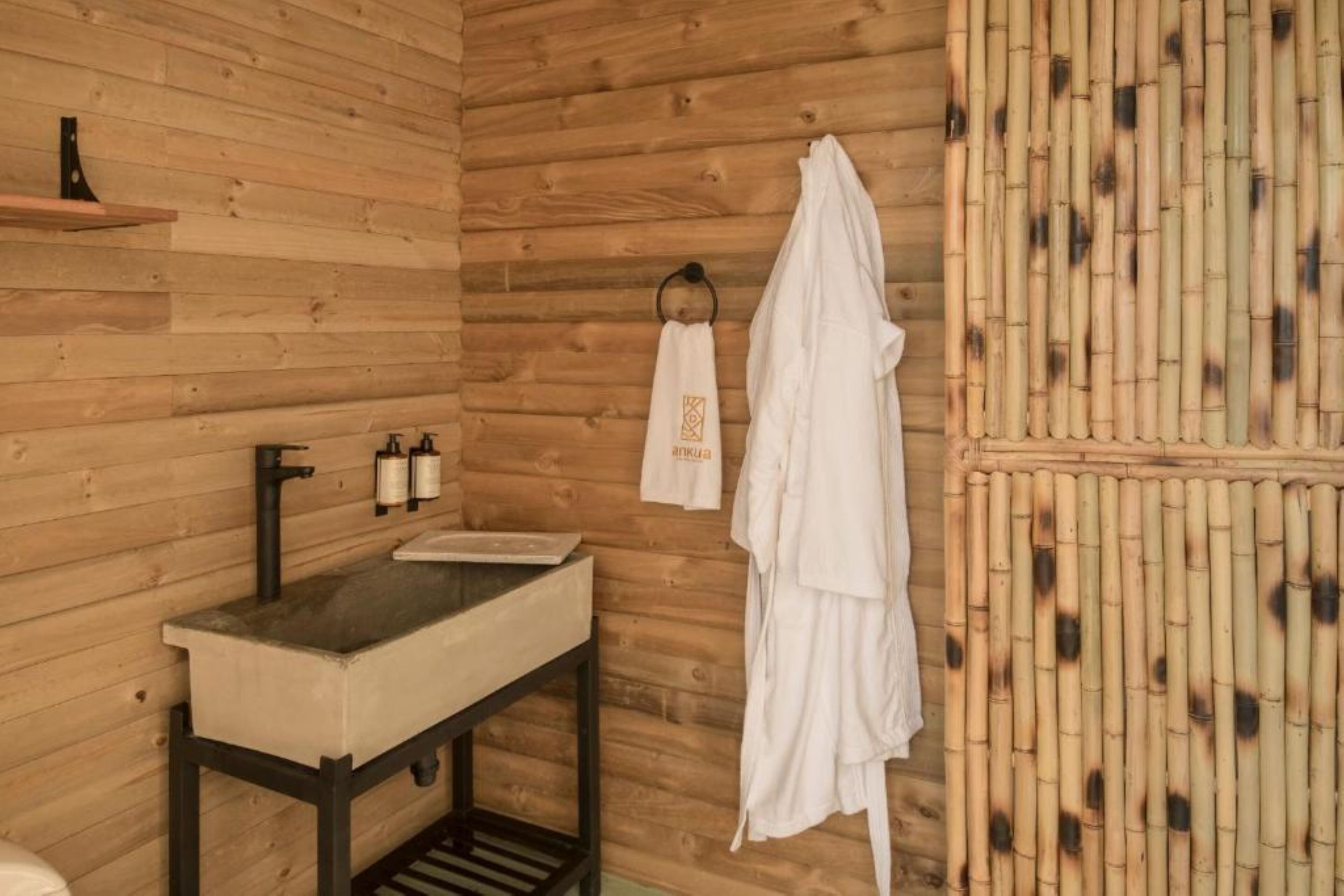 Baño de cabaña de madera con lavamanos, perchero para toallas y pared de bambú.