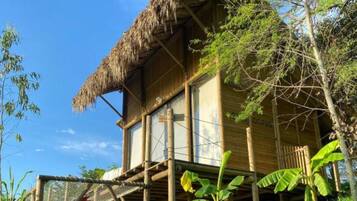 A wooden house with a thatched roof, elevated on stilts, surrounded by lush greenery.