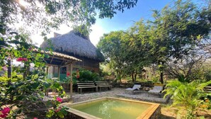 Una construcción con techo de paja y piscina, rodeada de vegetación y zonas de estar.