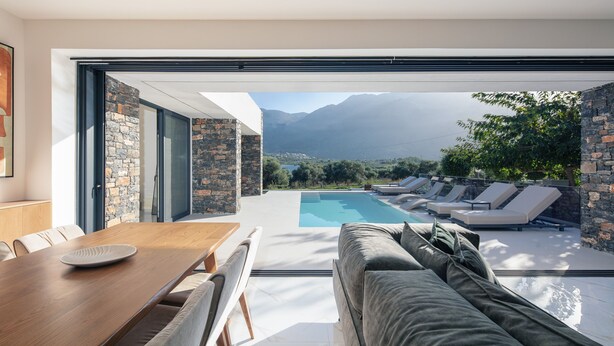 A modern dining area with a wooden table and chairs, a stone wall, and a view of a swimming pool and mountains. - Luxurious villa with a large pool, 750m from the beach. (Asprouliani)