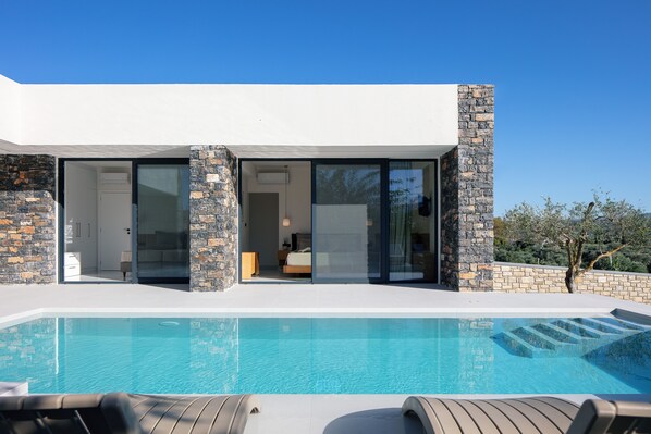 A modern house with a swimming pool, stone walls, and a bedroom visible through glass doors. - Luxurious villa with a large pool, 750m from the beach. (Asprouliani)