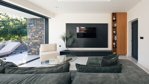 A modern living room with a large flat-screen TV mounted on the wall, a glass coffee table, and a dark green sofa. - Luxurious villa with a large pool, 750m from the beach. (Asprouliani)
