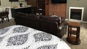 Een woonkamer met een donkerbruine leren bank, een geborduurde plaid, een klein houten bijzettafeltje, een flatscreen televisie aan een bakstenen muur en een open haard.