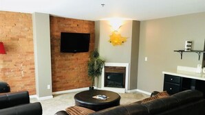 Sala de estar com parede de tijolos, TV de tela plana, lareira e área de jantar com balcão e prateleiras.
