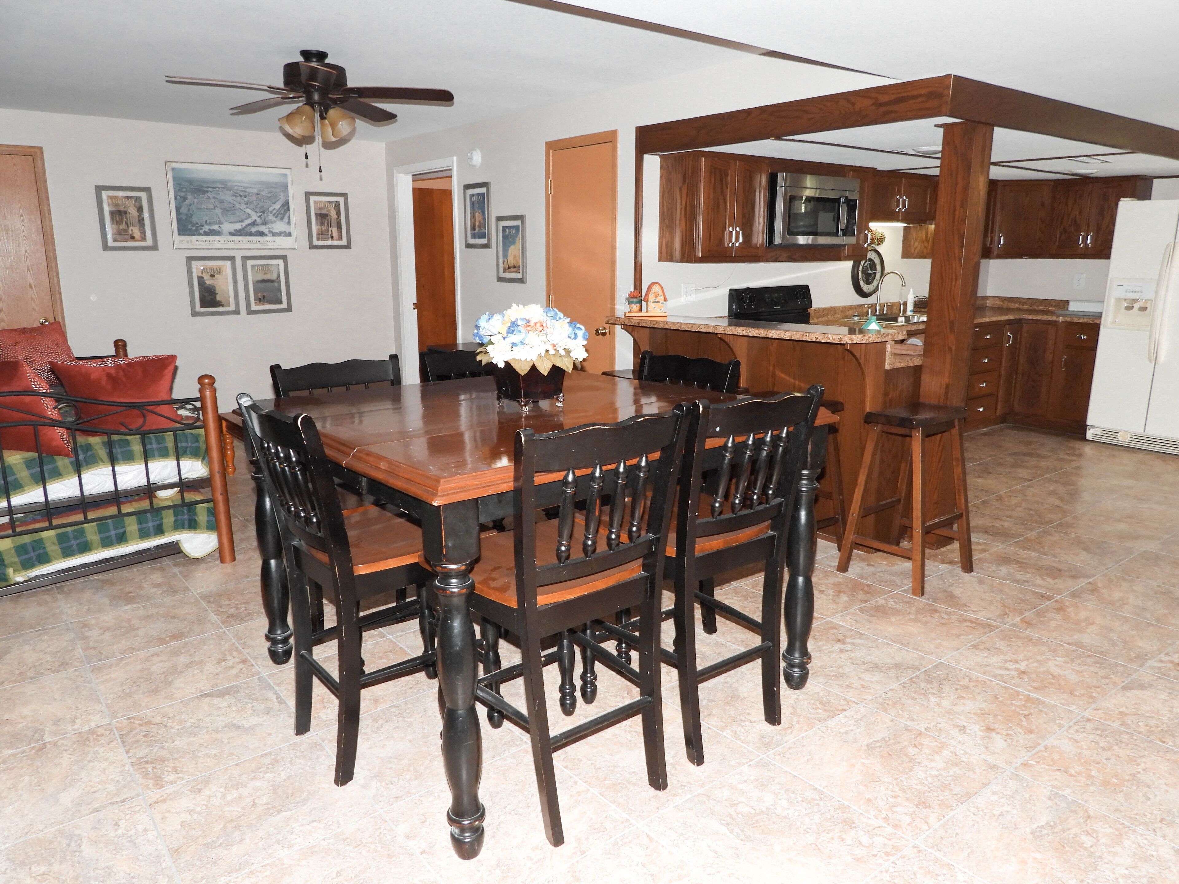 A dining area with a wooden table and chairs, a kitchen with wooden cabinets and a microwave, and a ceiling fan.