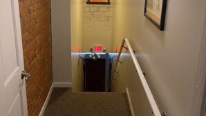 A narrow hallway with a brick wall on one side, a door at the end, and a staircase leading to an upper floor. There is a framed picture on the wall and a light fixture above the staircase. - Double Queen Guestroom (Corning)