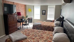 A hotel room with two beds, a desk, a dresser, a TV, and a brick wall. - Double Queen Guestroom (Corning)