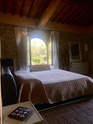 A bedroom with a large bed, a desk with a board game, a window with curtains, and a stone wall. - Room in B&B - Room Overlooking the Vineyards and Florence (Carmignano)