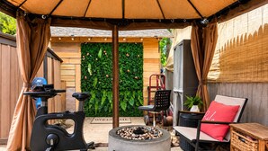 Un espacio al aire libre con unísones estacionaria, una mesa redonda con un brasero, una silla con cojín rojo y un mueble de almacenamiento de mimbre.