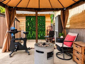An outdoor patio area with a stationary exercise bike, a round table with a fire pit, a chair with a red cushion, and a wicker storage unit. - Room-black-white Rm By Yale U (New Haven)