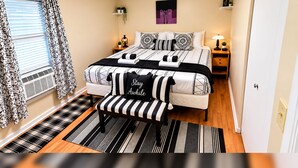 Ein Schlafzimmer mit einem Bett, Nachttischen, einer Bank, einem Fenster mit Vorhängen, einem an der Wand befestigten Regal und einer Tür.