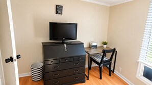 Ein Zimmer mit Fernseher auf einer Kommode, einem Schreibtisch mit Stuhl, einer kleinen Topfpflanze und einem Fenster mit Jalousien.