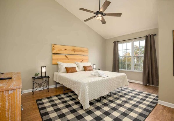 A bedroom with a large bed, a ceiling fan, a window with curtains, and a checkered rug.