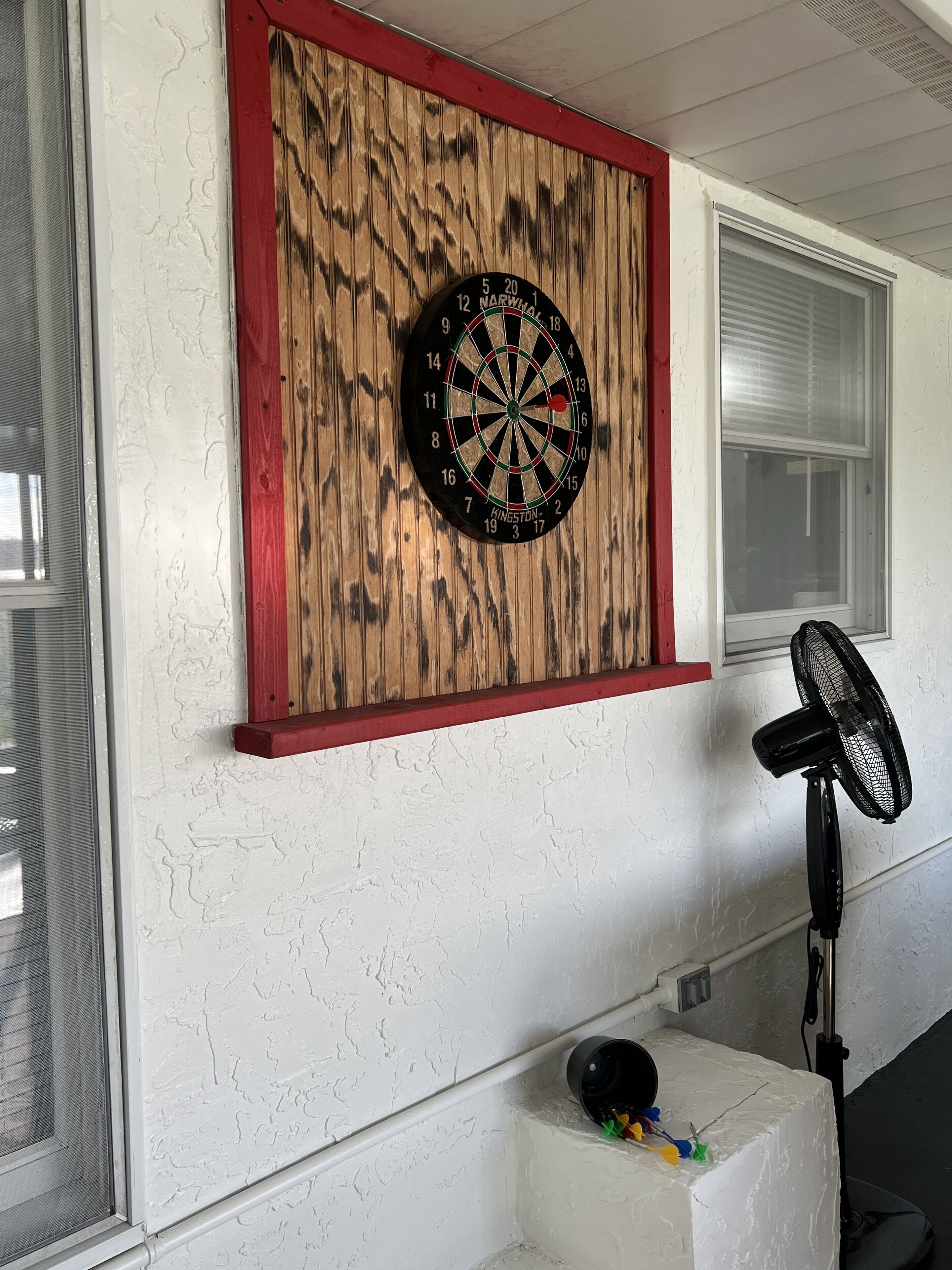 A dartboard mounted on a textured wall with a red frame, a window with blinds, and a standing fan.