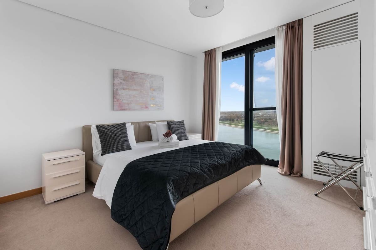 A bedroom with a bed, a bedside table, a window with a view, and a wall-mounted artwork.