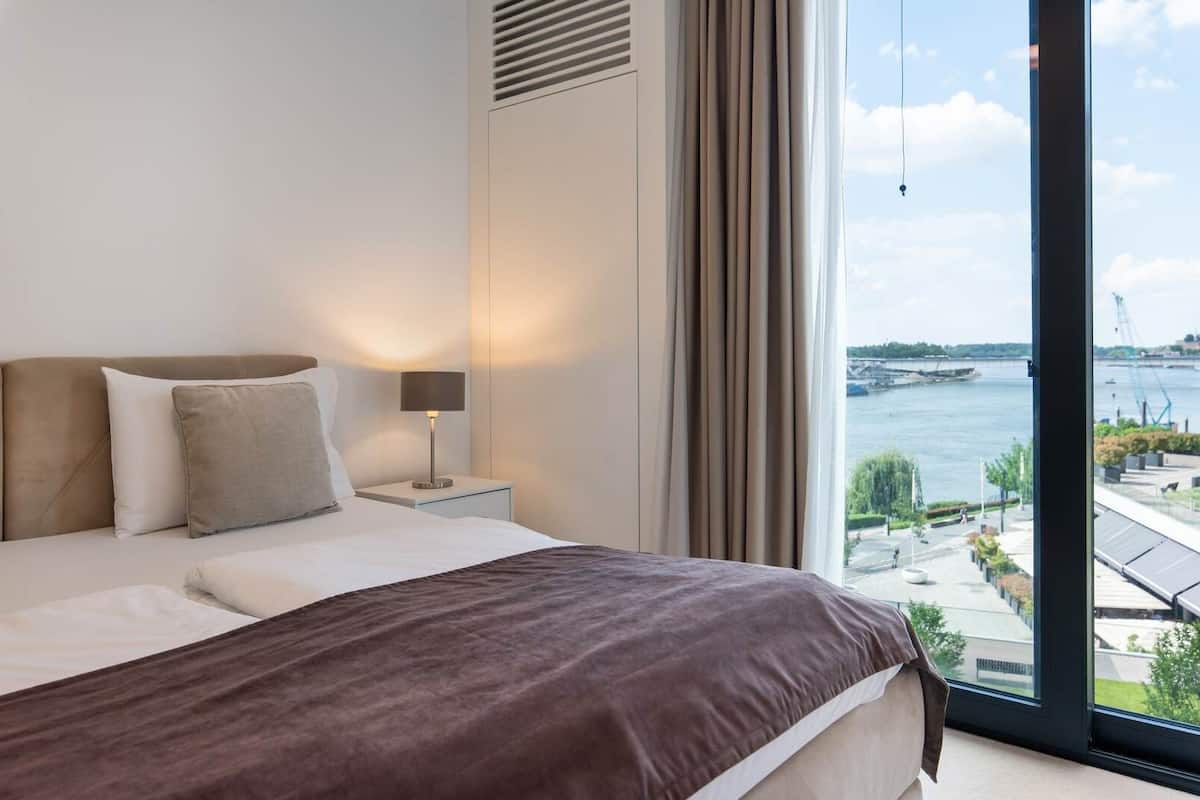 A hotel room with a large bed, a bedside table with a lamp, a view of a river and bridge, and a modern architectural design.