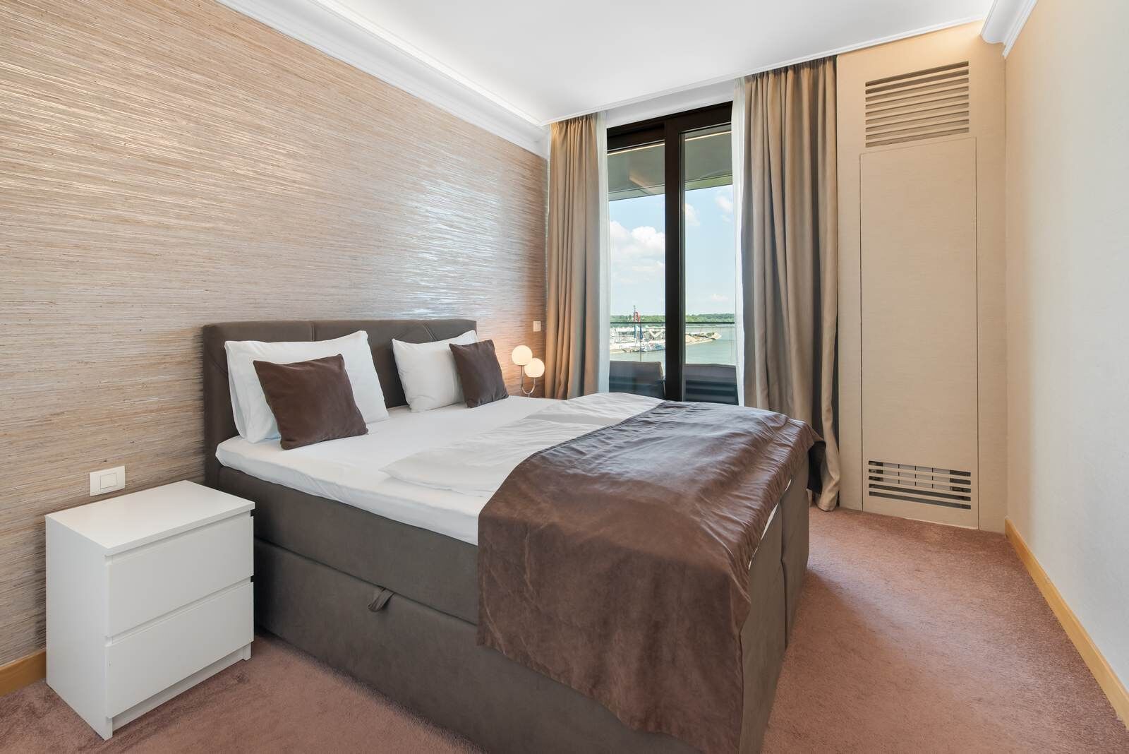 Una habitación de hotel con cama, mesita de noche, ventana con cortinas y vista al mar.