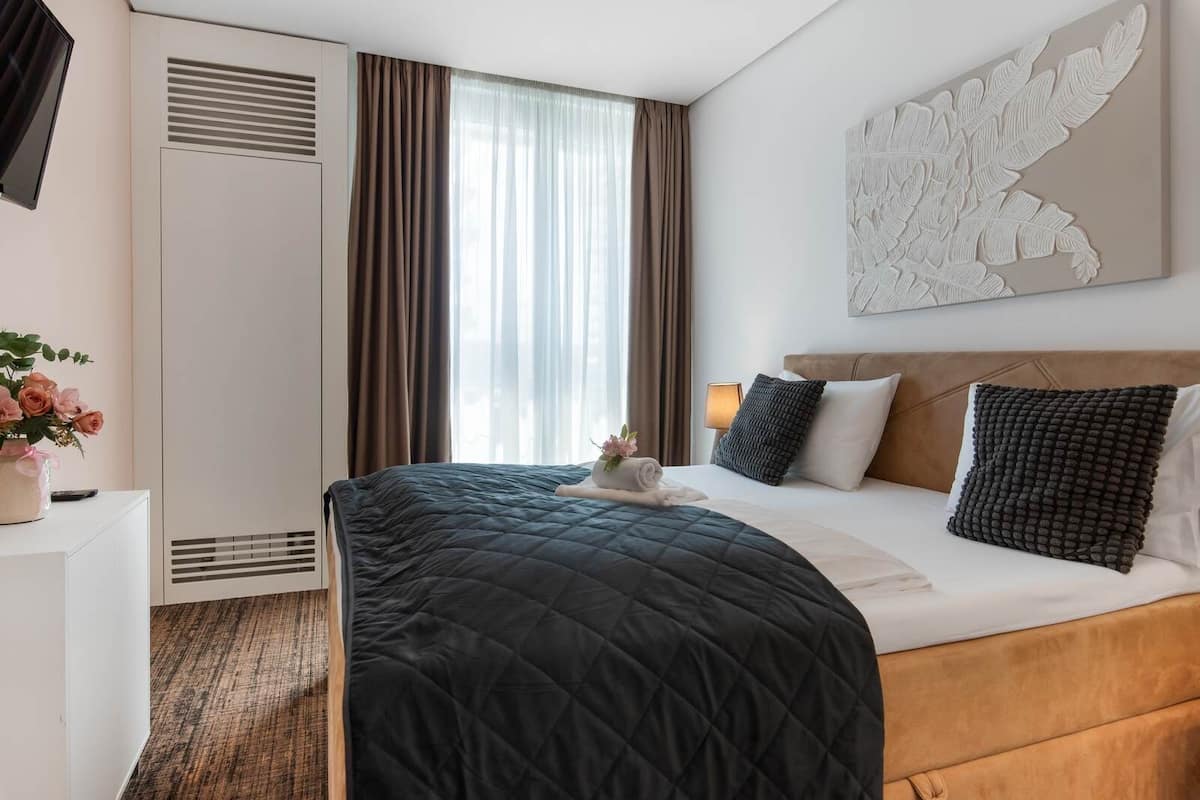A hotel room with a bed, a bedside table with a vase of flowers, a wall-mounted artwork, and a window with curtains.