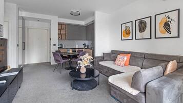 Un salon moderne avec un canapé gris, une table basse ronde et des fauteuils violets. On aperçoit une cuisine au fond, avec une table à manger et des chaises.