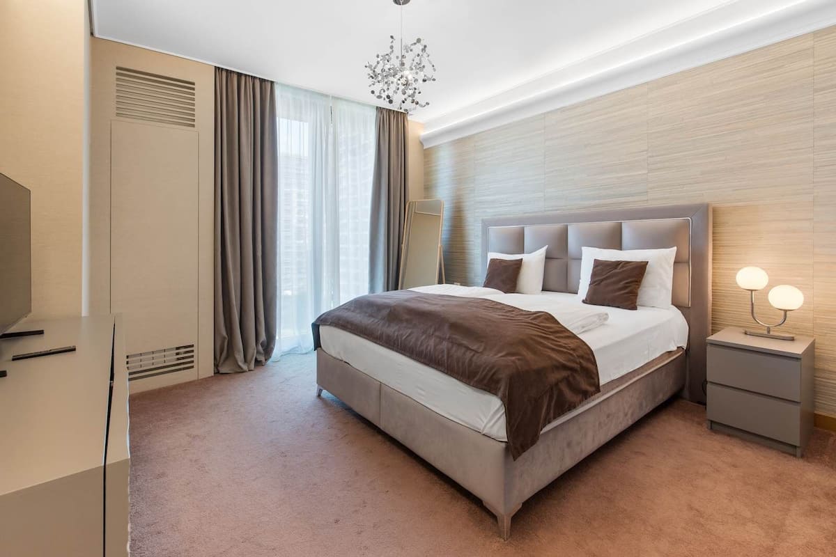 A modern hotel room with a large bed, a bedside table with a lamp, a wardrobe, and a large window with curtains.