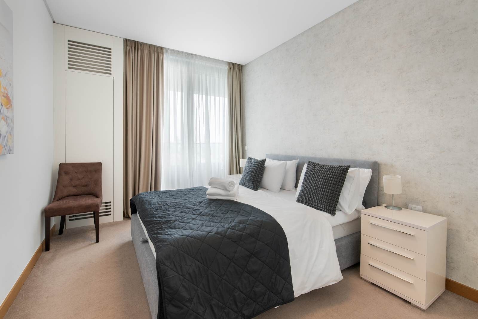 Una camera d'albergo con un letto, una sedia, un comodino e una finestra con tende.